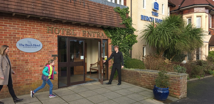 young man opening the front door of the hotel welcoming a running girl and her mum